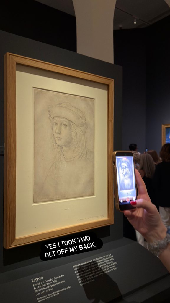 The same silverpoint self-portrait drawing by Raphael displayed in a wooden frame on a dark wall, with a wall label partially visible below. A visitor photographs it with a smartphone.