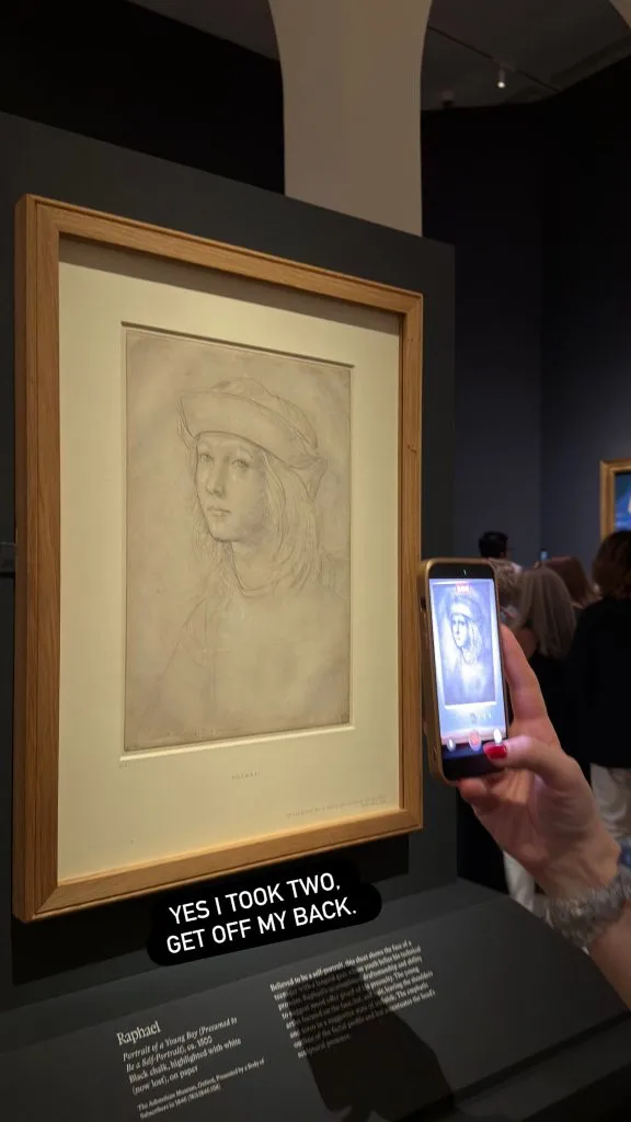 The same silverpoint self-portrait drawing by Raphael displayed in a wooden frame on a dark wall, with a wall label partially visible below. A visitor photographs it with a smartphone.