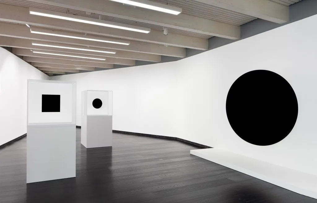 small black artworks are mounted on white podiums in a white walled space, on the right a black circle hovers on the wall