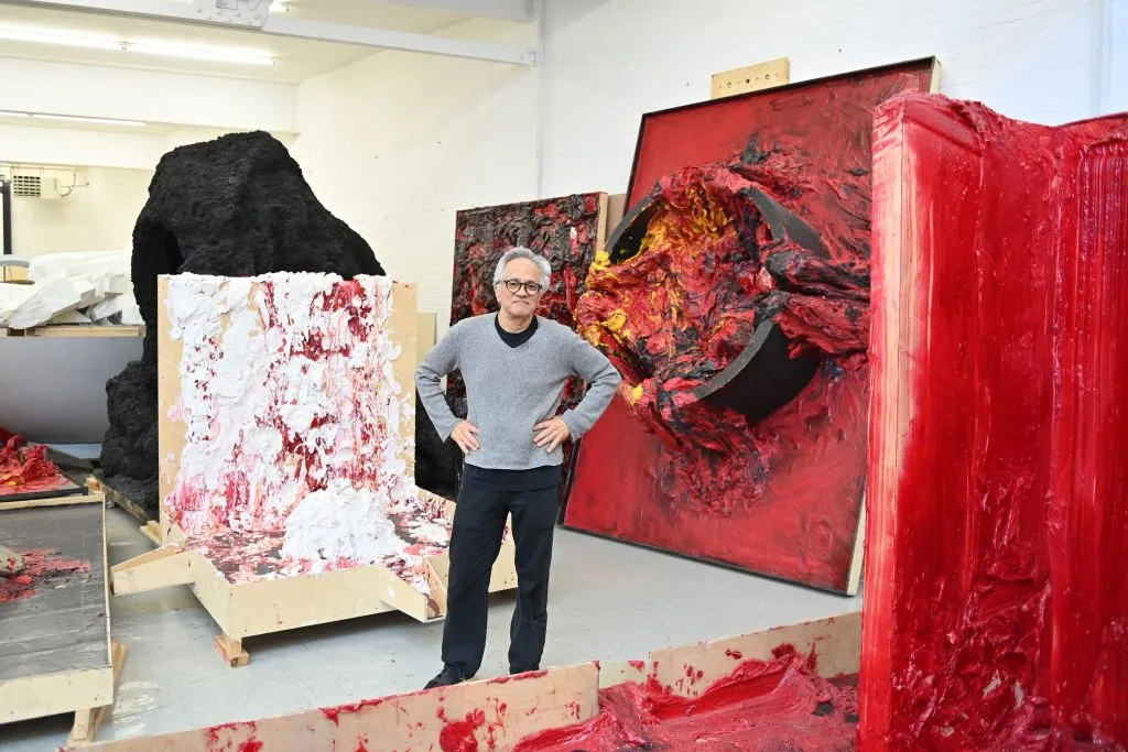Anish Kapoor stands in his studio surrounded by red pigment and wax sculptures in progress