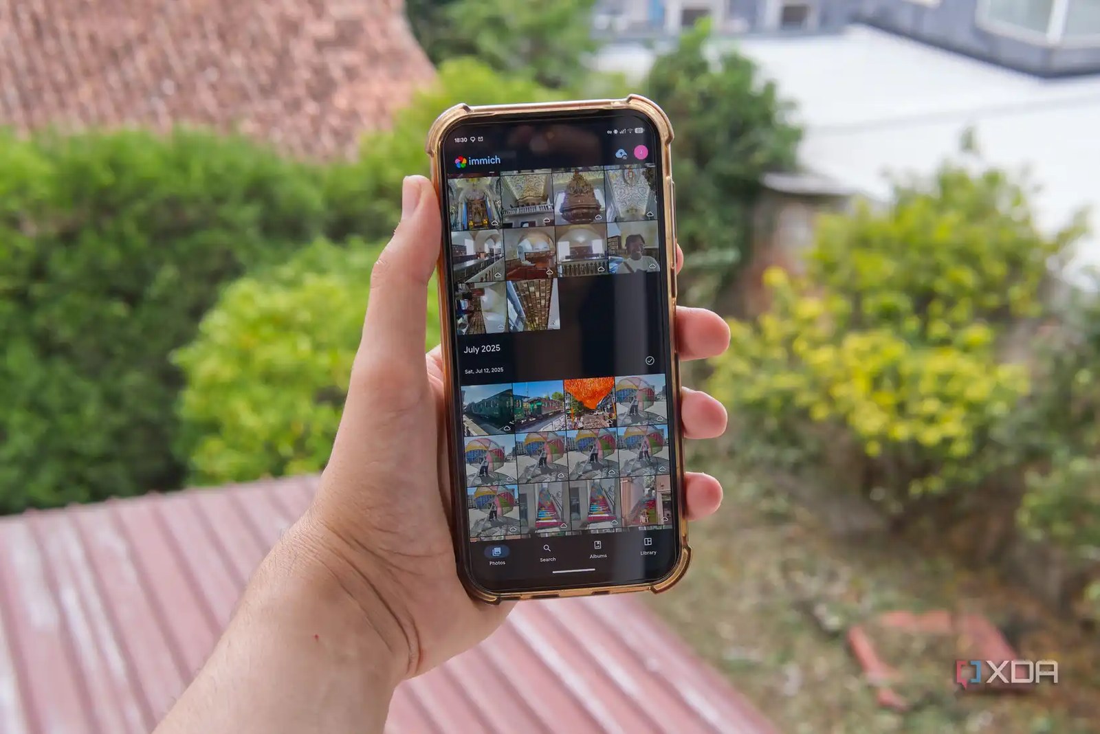 A phone held in a person's hand showing the Immich app, with trees and buildings in the background