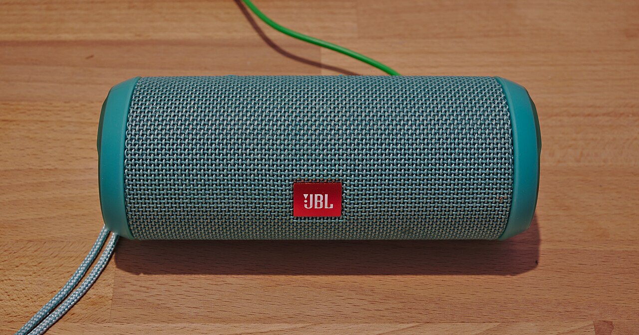 A JBL Flip 3 battery-powered and waterproof Bluetooth speaker connected to a charging cable. The length is ca. 17 cm, diameter 6.4 cm, weight 450 g.